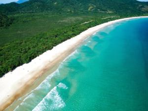 Foto panorâmica de uma enseada de águas límpidas verde-azuladas, praia de areia bem clarinha, seguida de floresta litorânea densa, com montanhas ao fundo