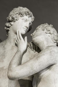 Foto detalhada da estátua de um casal: uma pessoa se apoiando delicadamente sobre o ombro da outra, com a mão tocando levemente sua face, e as duas se olhando apaixonadamente