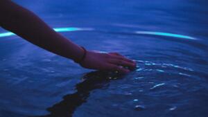 Foto detalhada do braço de uma pessoa utilizando uma pulseira vermelha discreta, estendendo a mão tocando levemente a superfície de um corpo de água